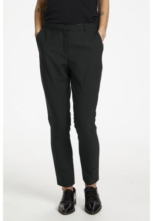 Person wears slim black trousers with side pockets and black lace-up dress shoes, standing against a white background.