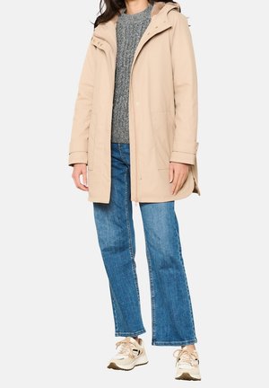 Parka beige avec capuche en fourrure, fermetures éclair et à pression, poches avant, associée à un pull gris et un jean évasé, portée avec des baskets.