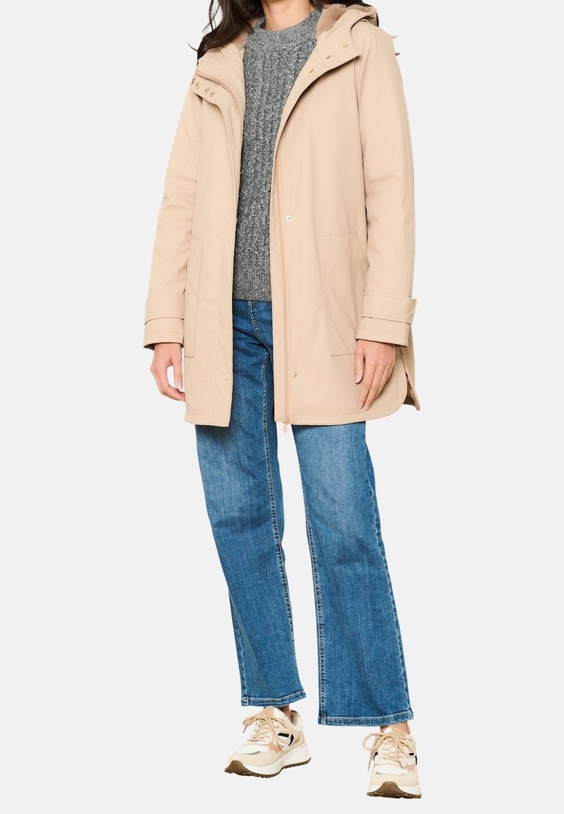 Beżowa parka z futrzanym kapturem, zapinana na zamek i napy, z przednimi kieszeniami, noszona z szarym swetrem i dzwonami, w połączeniu z trampkami.