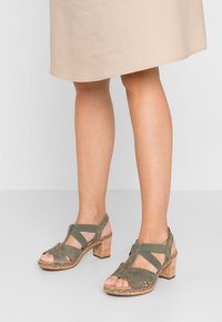 Jalat yang mengenakan rok khaki sepanjang lutut dan sandal block heel terbuka berwarna hijau suede dengan hiasan anyaman di latar belakang terang.