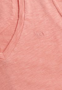 Chemise rose pâle à col en V avec un logo brodé discret sur la poitrine, montrant la texture du tissu et les détails des coutures.