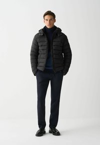 Črna pufasta jakna s horizontalnim prešitim vzorcem, z zadrgo in kapuco, v kombinaciji z mornarsko modrim puloverjem z visokim ovratnikom in temnimi hlačami, ki jih nosi model.