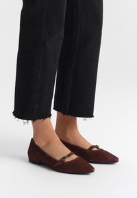Ballerines en daim marron avec un bout pointu et une seule sangle dotée d'une boucle en métal. Associées à un pantalon noir à ourlet effiloché.