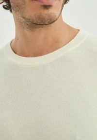 Pull en tricot crème clair avec un col rond, présentant un motif texturé et des poignets côtelés, montrant des détails fins dans le tissu.