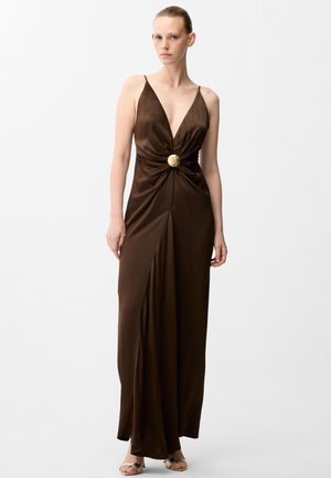 Femme portant une robe en satin marron sans manches avec un profond décolleté en V, une boucle circulaire dorée à la taille et une fente à l'avant, debout devant un fond blanc uni.