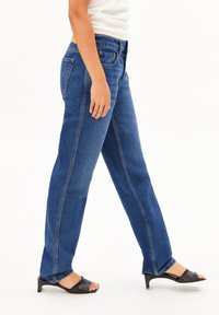 Jean en denim bleu avec une coupe droite, doté d'une taille haute et de coutures latérales visibles. Porté avec des sandales à talons noires.