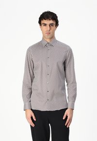 Chemise à manches longues boutonnée blanche avec un motif en losanges rouge et marine. Dispose d'un col pointu et d'une coupe ajustée.