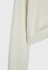 Manche de pull en maille écru et détail de bord-côte sur un fond clair uni.