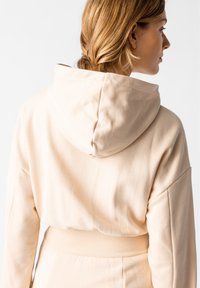 Beige verkürzter Hoodie mit großer Kapuze, aus weichem Material gefertigt. Verfügt über überschnittene Schultern und einen gerippten Saum. Glatte Textur.