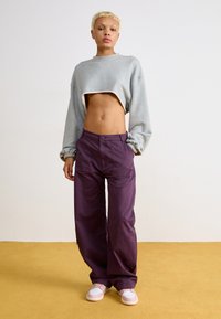 Sweatshirt gris court avec des poignets élastiques, pantalon violet à jambes larges avec poches avant, et baskets rose clair sur un fond orange-jaune.