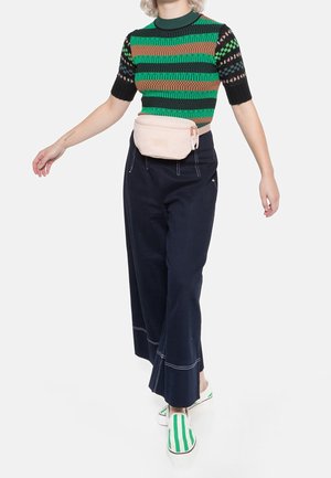 Persona che indossa un maglione a maniche corte a righe verdi e marroni, pantaloni larghi blu navy, scarpe slip-on a righe verdi e una marsupio beige, che cammina in avanti.