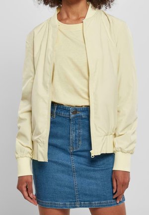 Nainen, jolla on päällään vaaleankeltainen vetoketjullinen takki, vastaava keltainen t-paita ja sininen farkkuhame, jossa on nappikiinnitys ja vetoketju.