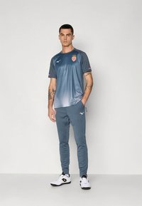 Mėlynas sportinis marškinėlis su logotipu ir gradientiniu raštu, derinamas su siaurėjomis kelnėmis. Komplektą papildo balti sportbačiai.