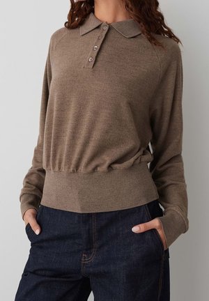 Le modèle porte un haut en maille taupe à manches longues, avec un col et une patte de boutonnage, ainsi qu’un jean indigo foncé. Il se tient debout, les mains dans les poches.