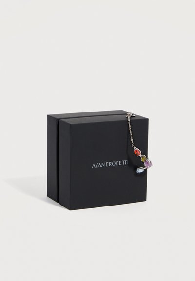 Scatola regalo nera con logo argentato e braccialetto a catena argentato con pietre preziose multicolori appoggiato sul bordo.