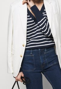 Blazer blanc avec boutons dorés porté sur un pull en maille rayé bleu marine. Jean taille haute bleu foncé, maintenu par une ceinture noire.