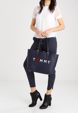 Sac cabas bleu marine avec le lettrage blanc « TOMMY » et un cœur rouge. Fabriqué en matériau lisse, avec des anses et une forme structurée.