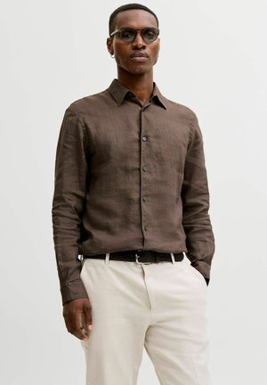 Mand iført mørkebrun langærmet skjorte med knapper, beige bukser, sort bælte, runde solbriller og en lille ørestik, står med den ene hånd i lommen.