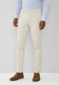 Světle béžové kalhoty vyrobené z hladké tkaniny, slim fit, s předním zapínáním na knoflík, poutky na opasek a bočními kapsami, doplněné o hnědé semišové boty.