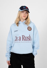 Jeune personne aux cheveux blonds portant un sweat à capuche bleu clair avec le texte « In a Rush International » et une casquette marine avec un logo rouge et bleu.