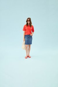 Femme en t-shirt rouge, jupe en jean, tongs rouges et lunettes de soleil tenant un sac filet beige, debout devant un fond bleu clair uni.