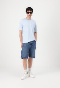 Light blue short-sleeve T-shirt paired with dark denim shorts. The outfit is completed with white sneakers and black sunglasses.