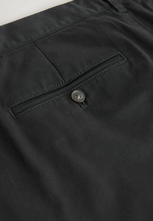 Primer plano de pantalones de tela negra con un bolsillo trasero de vivo asegurado con un solo botón negro.