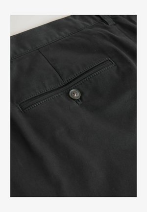 Gros plan d’un pantalon en tissu noir avec une poche passepoilée à l’arrière maintenue par un seul bouton noir.