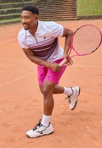 Polo shirt rayé blanc et rose, shorts roses, et baskets blanches avec des accents noirs. Un joueur masculin tient une raquette de tennis rose sur un court en terre battue.