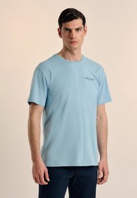 Camiseta de algodón azul claro con mangas cortas, cuello redondo y un pequeño logo impreso en el lado izquierdo del pecho. Textura suave, ajuste casual.