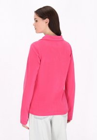 Pullover in pile di colore rosa brillante, con una texture morbida, maniche lunghe e colletto ripiegato, abbinato a jeans azzurri chiari.