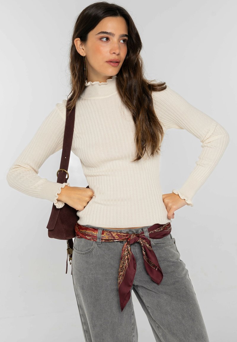 Suéter de cuello alto acanalado color crema con puños fruncidos, combinado con pantalones grises de talle alto y una bufanda burdeos con estampado en la cintura.