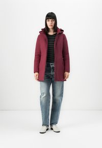 Vrouw in een kastanjebruine konijnenjack over een zwart gestreept shirt, gescheurde blauwe jeans en witte schoenen, staand tegen een effen witte achtergrond.