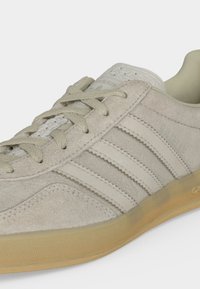 Μπεζ σουέντ αθλητικό παπούτσι Adidas με τρεις ρίγες, δεμένα κορδόνια και διάφανη καουτσούκ σόλα, εμφανίζεται σε κοντινό πλάνο σε ουδέτερο φόντο.