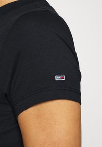 Svart t-shirt med kort ärm i bomull, med en liten broderad logotyp med röda, vita och blå detaljer på ärmen.