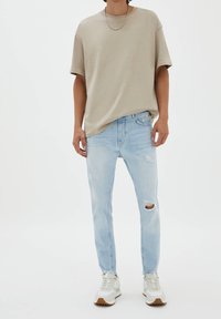 Camiseta beige de gran tamaño, vaqueros ligeros desgastados de color azul, zapatillas blancas con acentos marrones, collar de cadena simple, colores neutros.