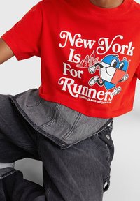 Červené zkrácené tričko s bílým nápisem "New York Is For Runners" a barevným srdíčkem, spárované s tmavými denimovými kalhotami.
