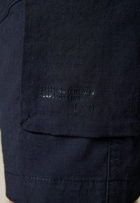 Shorts din țesătură texturată de culoare bleu marin, cu un buzunar proeminent ce prezintă detalii imprimate, inclusiv "ADAPT FOR LIVING" și alte simboluri.