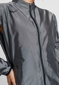 Nike zipová bunda v tmavě šedé barvě. Má hladký, lesklý povrch, vysoký límec, zapínání na zip a boční kapsy s elastickými manžetami.