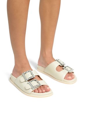 Sandali scivolosi di colore crema realizzati in materiale sintetico liscio, caratterizzati da due ampie fibbie con strass e un design con punta aperta.