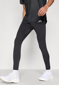 Leggings deportivos negros con un diseño ajustado, que presentan costuras decorativas blancas y un pequeño logo en la pretina, combinados con zapatillas blancas.