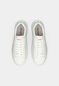 Zapatillas deportivas blancas de cuero con punta redonda, cordones planos y el logotipo "HUGO" en rojo en la lengüeta y los lados. Textura suave, diseño minimalista.