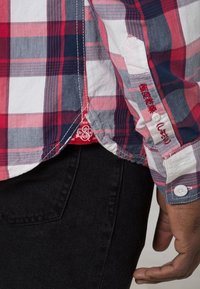 Gros plan d'une personne portant une chemise à carreaux rouge, blanche et bleue avec un texte japonais sur le poignet de la manche, associée à un pantalon noir.
