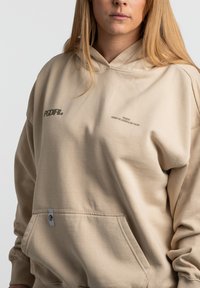 Beige Kapuzenpullover aus weichem Stoff, mit einer Kängurutasche, gesticktem Logo und Textdetails auf der Brust.