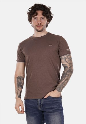 T-shirt marrone a maniche corte con colletto rotondo, realizzato in materiale morbido. Presenta un logo sul petto e del testo sulla manica. Modello lo indossa con dei jeans.