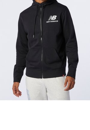 Zwarte zip-up hoodie met New Balance-logo op de borst, gedragen door een persoon, gecombineerd met lichtgrijze joggingbroek met blauwe tekst op de dij.
