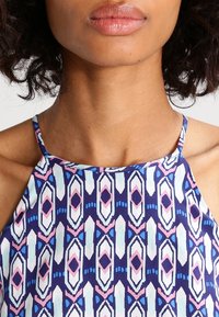 Nära bild av en kvinna som bär en ärmlös topp med blå, rosa och vita geometriska mönster och tunna axelband.