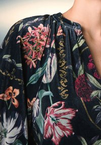 Tessuto nero con motivi floreali colorati e dettagli intricati, con scollo arricciato. La texture appare morbida e ricca.