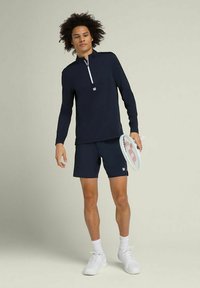 Top sportivo a maniche lunghe blu navy con mezza zip e tasca frontale, abbinato a pantaloni corti blu navy. Il modello tiene una racchetta da tennis. Sneakers e calze bianche.