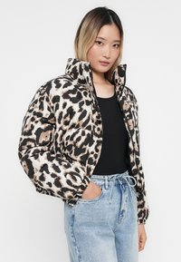 Een pufferjack met een luipaardprint in beige, zwart en bruine tinten. Heeft een hoge kraag en elastische manchetten. Gemaakt in combinatie met lichte spijkerbroeken.